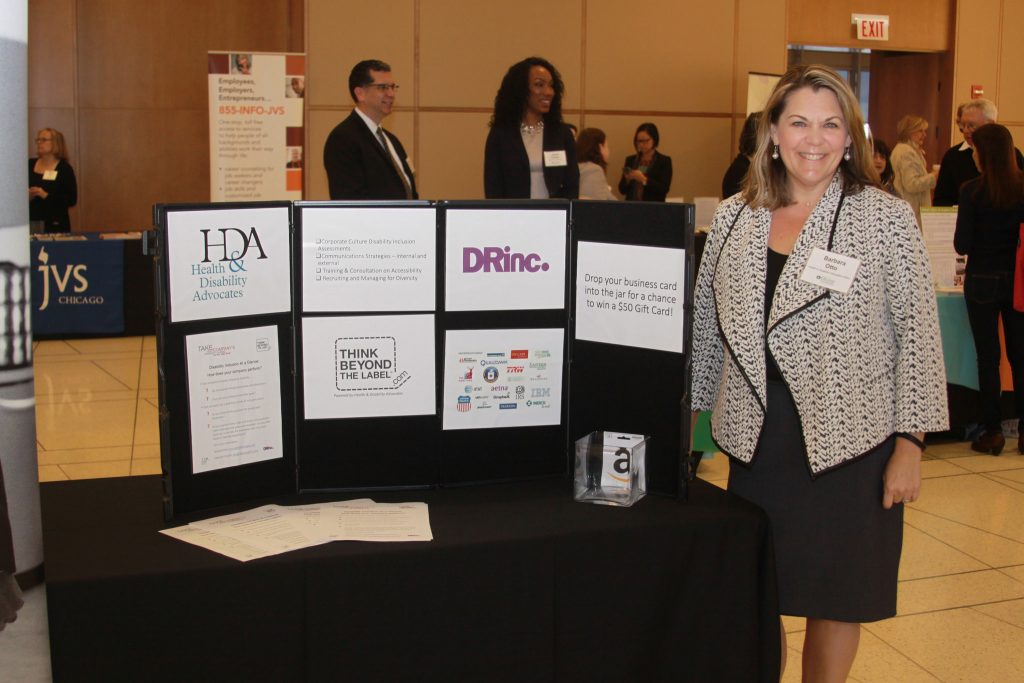 Barbara Otto stands at the Health & Disability Advocates exhibitor table at the Disability Inclusion Opportunity Summit