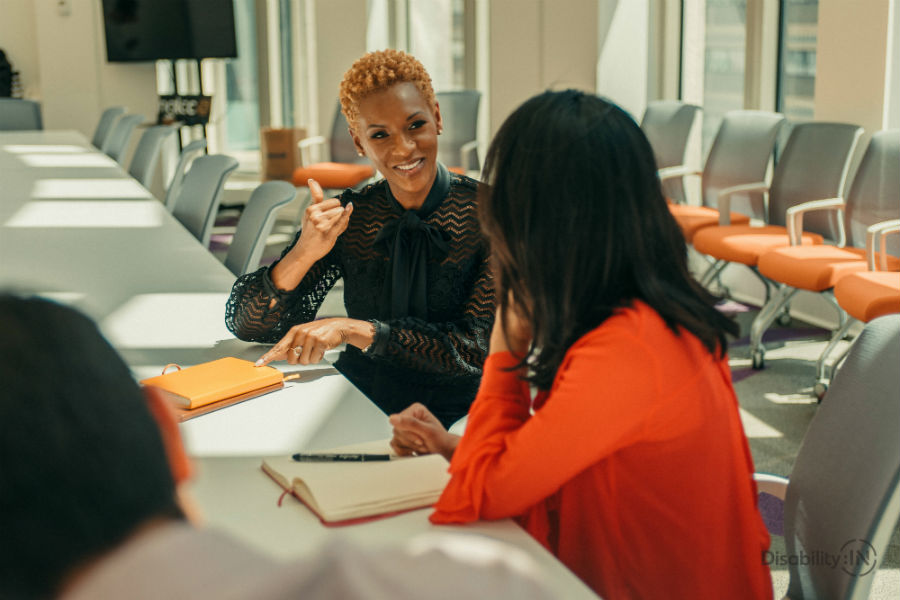 Two women sitting at a board table, one person is smiling and signing ASL.